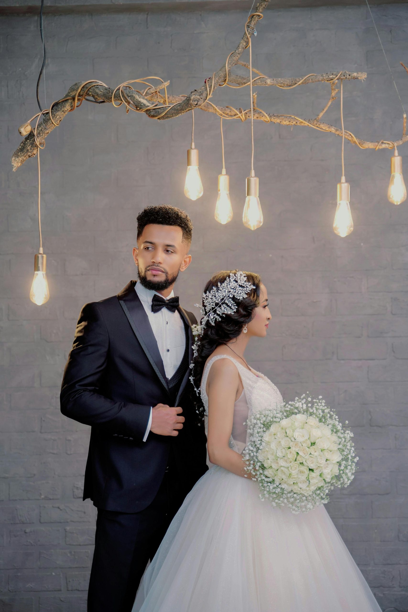Elegant bride and groom in wedding attire posing with bouquet under decorative lights
